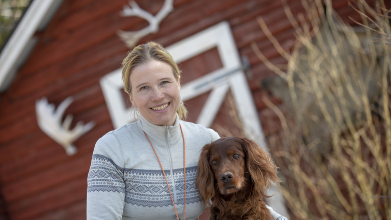 En kvinna med stickad tröja tillsammans med en brun hund framför en röd lada med älghorn på fasaden. 