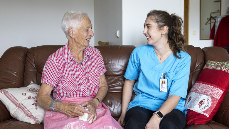 En äldre kvinna och en yngre undersköterska sitter i en soffa