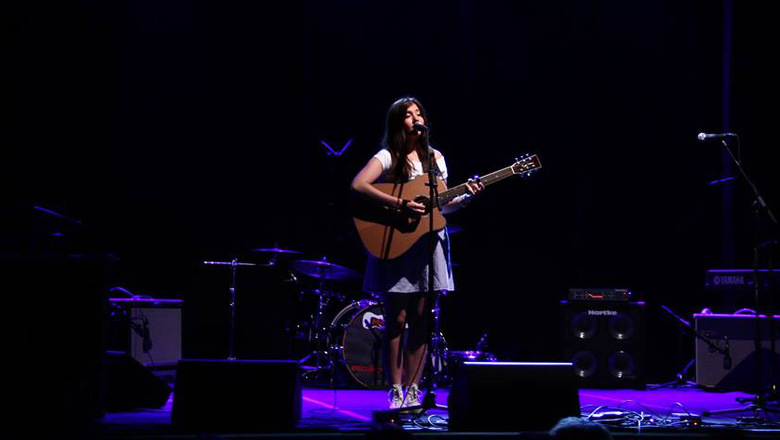 Bild på en tjej med gitarr som sjunger från en mörk scen med scenbelysning