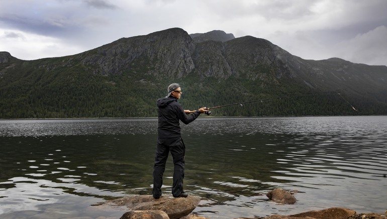 En man som fiskar vid sjö med fjäll i bakgrunden.