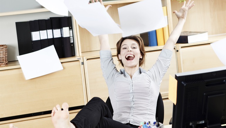 Bild på glad kvinna vid ett kontorsbord som kastar papper i luften