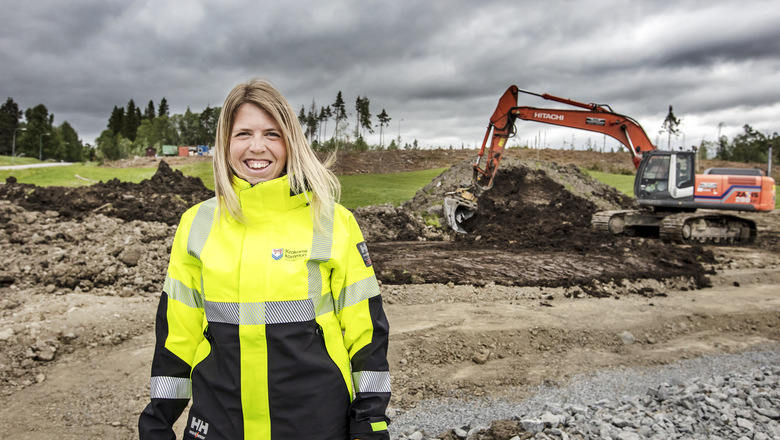 En leende kvinna i varselkläder står utomhus på en byggarbetsplats i bakgrunden syns en grävmaskin