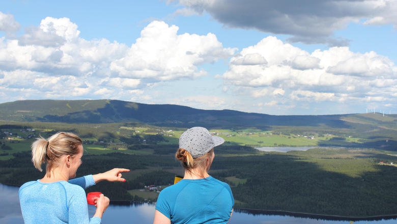 Två personer dricker kaffe och pekar på utsikten över sjö och böljande landskap från en höjd